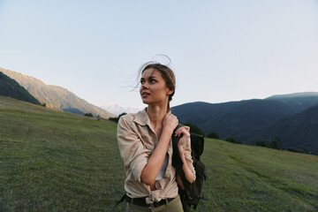 Fototapeta premium Young woman traveler stands on green mountain meadow with backpack looks away thoughtful enjoying nature and fresh air during hiking trip in clear sky daylight.