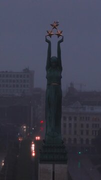 An aerial perspective captures the Freedom Monument in Riga, Latvia, standing above urban street traffic during foggy twilight conditions.