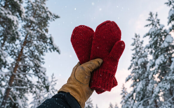 Person wearing leather glove holding red knitted mittens in a snowy winter forest