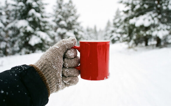 Gloved hand holding red coffee mug in snowy winter forest landscape for cozy seasonal concept