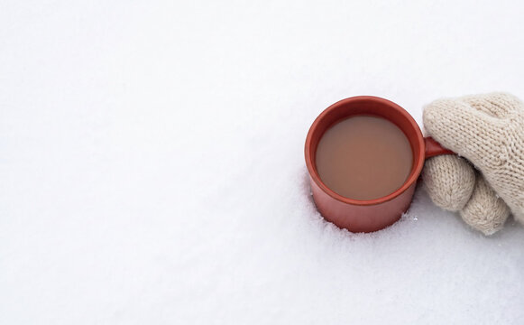 A hand in a knitted mitten holding a warm hot drink in a red mug on a cold white snow background representing winter comfort and relaxation