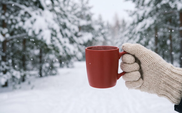A person in a knitted mitten holding a hot red coffee mug in a snowy winter forest