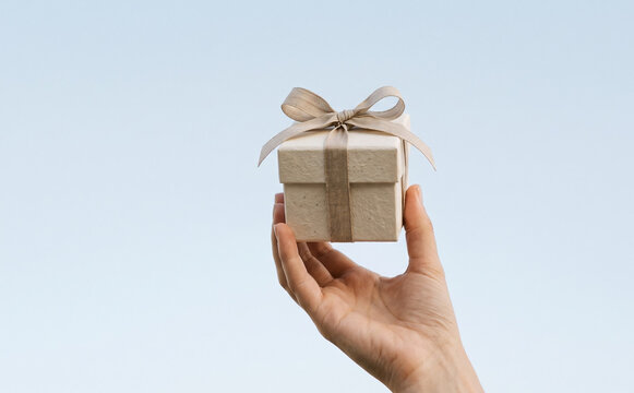 Hand holding a small gift box with a ribbon against a light blue sky representing celebration and surprise