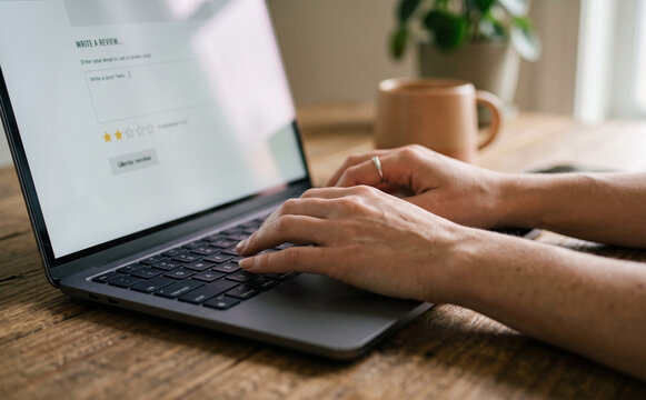 Close-up of a Caucasian woman writing a product review online using a laptop computer at a wooden table