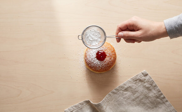Person sifting powdered sugar over a jelly donut on a wooden table, top view concept for baking and pastry preparation