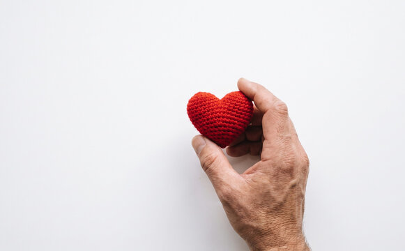 Close up of an adult male hand holding a small red crocheted heart on a white background representing love and charity