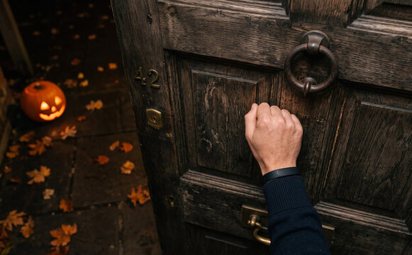 A person knocking on a rustic wooden front door of house number forty two with a glowing jack o lantern on the porch