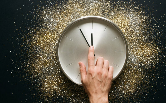 A person setting a minimalist metallic clock surrounded by sparkling gold glitter dust on a dark background