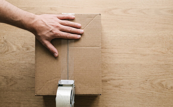 Top view of a person sealing a cardboard box with adhesive tape on a wooden table, representing shipping and packaging