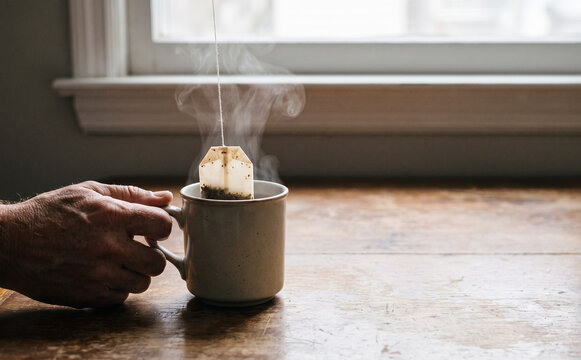 Steaming hot cup of tea with teabag on a wooden table being held by an adult man