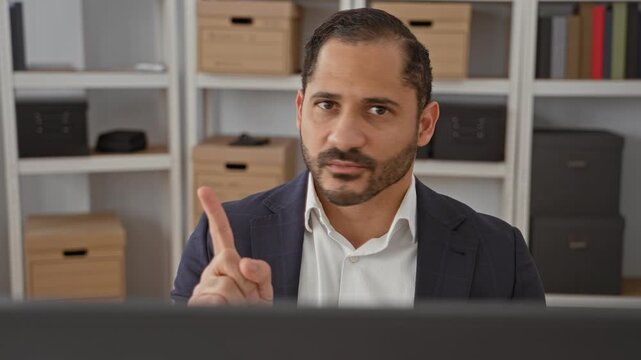 Man in suit at computer pointing finger toward camera with raised index near monitor and storage boxes on shelves in building; assertive focus.