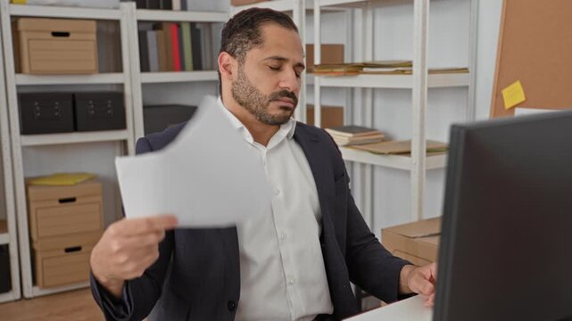 Man holding and fanning paperwork with one hand while seated at a desk with a computer monitor and storage shelves in a building; work stress.