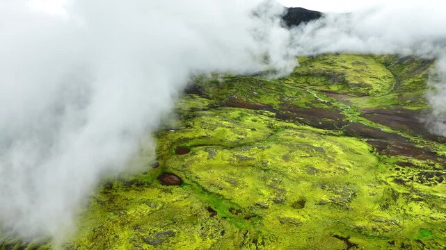 Oblique aerial reveal of moss-covered volcanic plateau as low clouds pull back to expose sinuous shallow stream and peat pools, with moss, stream, plateau, valley detail