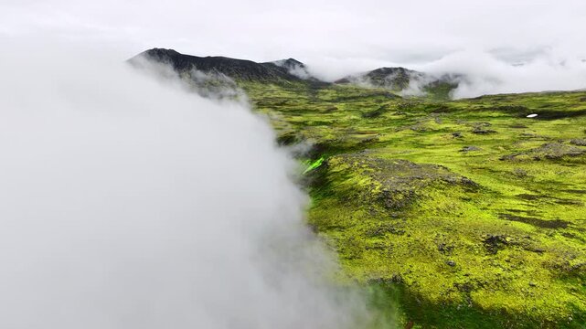 Oblique aerial reveal of moss-covered volcanic plateau as low clouds pull back to expose sinuous shallow stream and peat pools, with moss, stream, plateau, valley detail