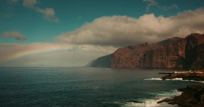 Ocean landscape with volcanic rocks and bright rainbow on cloudy sky. Los Gigantes cliffs in Tenerife.