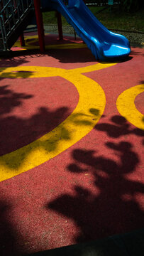 Colorful playground with red yellow rubber floor and blue slide. Tree shadows create abstract pattern. Concept of childhood, play and outdoor recreation.