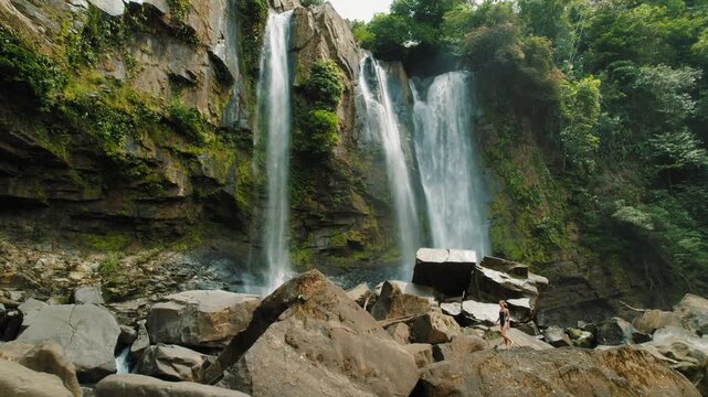 Woman walking on large rocks near a tall Nauyaca waterfall and tropical trees forming a lush natural paradise in Costa Rica