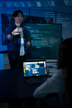 Smiling Asian male software developer gesturing during a presentation with programming code projected on a screen in a blue-lit room