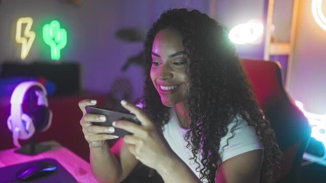 Woman holds smartphone with both hands and smiles, right fist clenched near chin while playing a mobile game in studio; joyful focus.