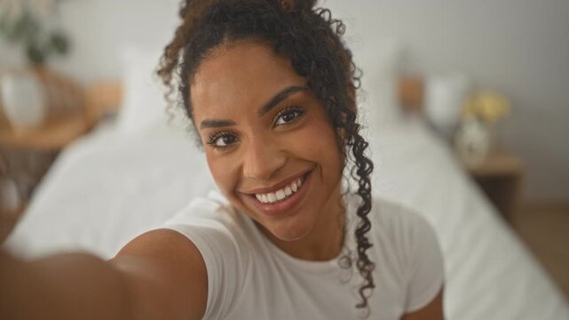 Woman on bed smiling with arm extended for selfie in building, curly hair and white t shirt, bedside plant and lamp visible; joyful connection.