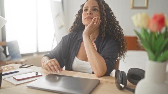 Woman resting hand on cheek at laptop on a desk in apartment with phone, notebook, headphones and tulips visible; quiet contemplation.