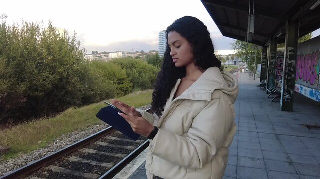 Beautiful mixed-race woman with long curly hair using a digital tablet while waiting for a train on the platform, commuting in the city with modern technology and public transportation