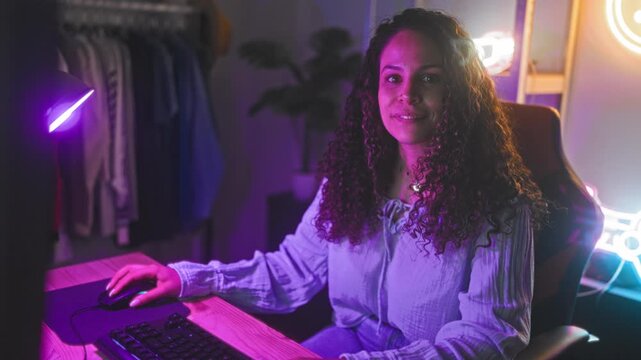 Woman with curly hair smiling, right hand on mouse and left near keyboard at gaming desk with neon controller sign in studio; joy connection.