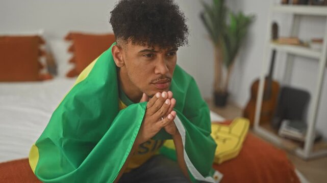 Man wrapped in brazil flag on bed with hands clasped praying and head bowed inside a building; despair team support.