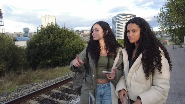 Two beautiful young twin sisters with curly hair smiling and waving goodbye while standing on a train station platform with their luggage, ready for a new journey and adventure together
