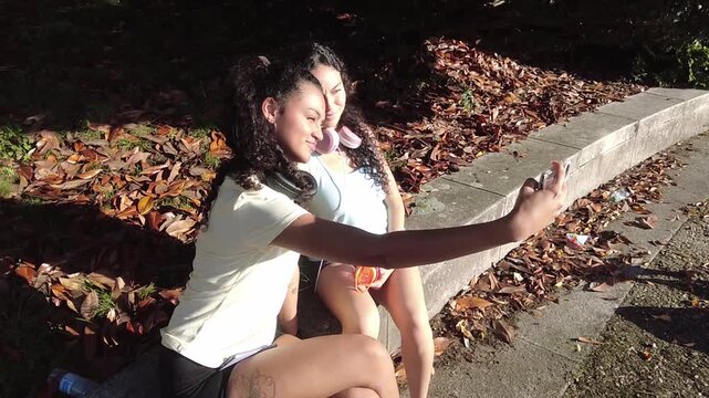 Two smiling young women in sportswear sitting on a curb in a park with autumn leaves, taking a selfie together with a smartphone and then looking at the picture, enjoying a sunny day