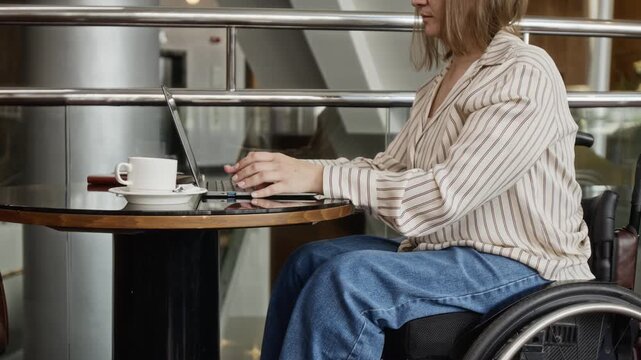 Young adult Caucasian woman with disability sitting in wheelchair and typing on laptop in cafe. Footage represents equal opportunities, remote employment, and digital accessibility concepts