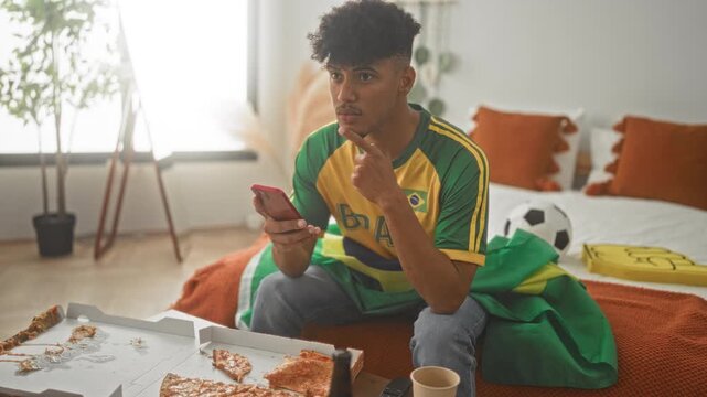 Man in brazil jersey holds smartphone and points while sitting on bed in bedroom; team spirit joy.