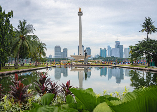 Jakarta, Indonesia - August 28, 2026: View of the National Monument, known as Monas, reflected in a pond with tropical plants and the Jakarta skyline in the background.


