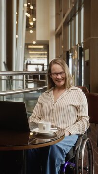 Portrait of Caucasian woman with disability sitting in wheelchair, typing on laptop in cafe and smiling at camera. Ideal for promoting workplace diversity and inclusion, vertical