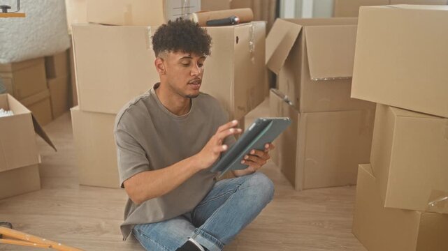 Young hispanic man tapping tablet while seated on floor amid stacked cardboard moving boxes and an orange work light in a building; focused transition.