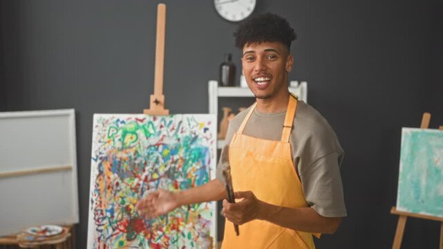 Man painter in orange apron holding paintbrushes and smiling while gesturing toward a colorful abstract canvas and easel in studio; creative joy.