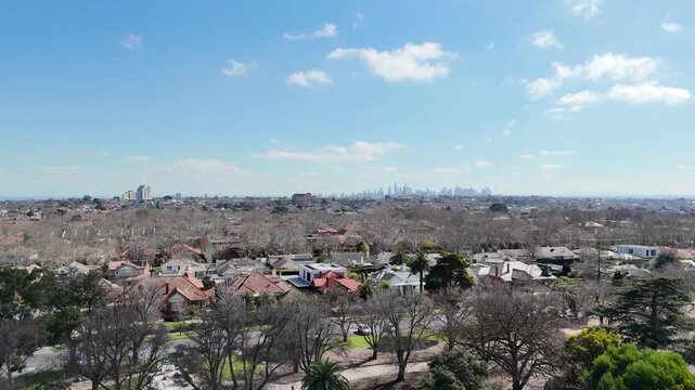 Aerial view over residential houses and bare winter trees taking off from a parkland with public green space in Malvern East, a suburb in Victoria, Australia, with the distant Melbourne city skyline. 