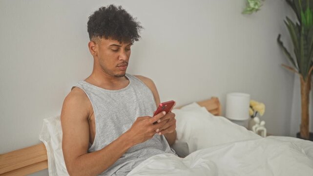 Man sitting on bed holding red smartphone and rubbing eyes with hand in a bedroom building; morning fatigue.