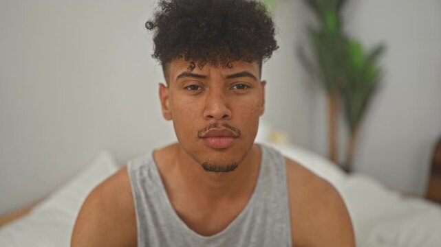 Man on bed with bare shoulders staring at camera in studio with grey tank top and potted plant by headboard; introspection solitude.