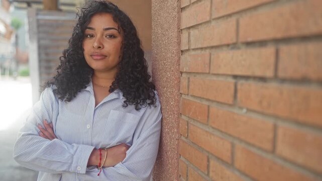 Woman with arms crossed leaning against a brick wall on a street, wearing striped shirt and bracelets; confidence warmth authenticity.