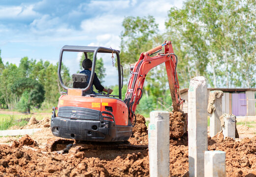 Compact orange mini excavator digging soil at a rural construction site during foundation preparation work on a sunny day.