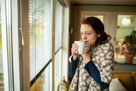 Young woman wrapped in blanket drinking tea by window at home