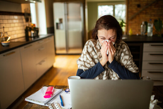 Ill woman blowing nose while working from home in kitchen