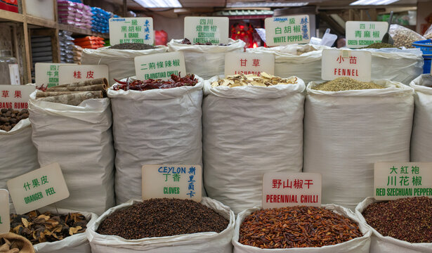  White sacks filled with dried spices, peppercorns, and chillies are displayed with bilingual labels at a market stall.