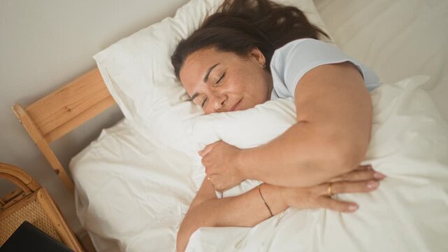 Woman hugging a pillow with closed eyes, face resting and forearm visible on a bed with white linens inside a building; serenity comfort.
