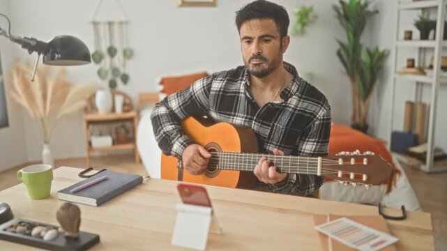 Man playing a classical guitar with visible hands and smartphone on table in bedroom, wearing plaid shirt, seated at desk; concentration.