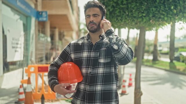 Man engineer holding orange hardhat with phone to ear speaking on smartphone on street; site inspection determination.
