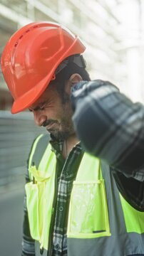 Man engineer in orange hardhat and reflective vest rubbing neck at a building site outdoors; workplace pain.