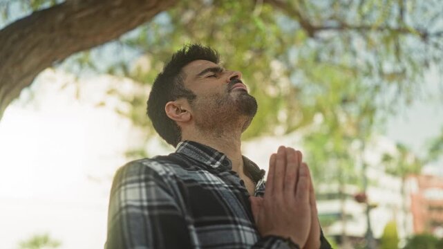 Man with hands pressed together in prayer, head tilted upward, eyes closed, wearing plaid shirt, standing beneath a tree on a street; serenity.
