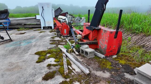 Unused agricultural snow blower and other forgotten junk resting on a cracked concrete slab at old farm